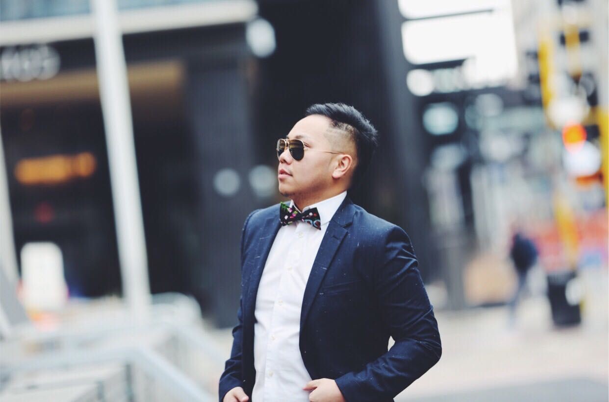 Man in a suit with a bow tie and sunglasses standing outdoors in an urban setting.